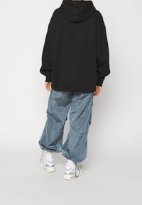 Sudadera negra con capucha de corte holgado, combinada con pantalones cargo azules brillantes. Calzado: zapatillas gris y blanco con suela gruesa.