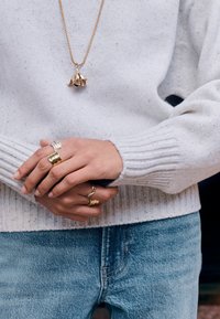 Gold pendant necklace on a person wearing a white ribbed sweater, showcasing multiple rings: silver, gold, and textured silver in varying designs.