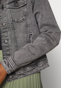 Grå denimjacka med knäppning fram, fickor med lock och ribbade ärmmuddar. Materialet har en mjuk textur med synliga sömnadsdetaljer.