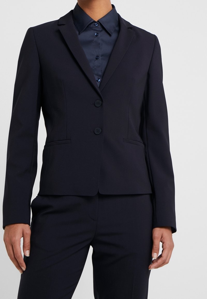 Námořnická modrá přizpůsobená blazer s jedním knoflíkem, s klopami a dvěma předními kapsami. Hladká tkanina s propracovaným střihem.