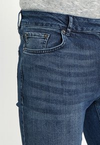 Jeans de mezclilla azul oscuro que cuentan con un bolsillo frontal, costuras visibles y una textura suave. La tela tiene un ligero efecto deslavado.
