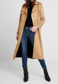 Abrigo de trench de suave ante en tono beige, con cuello de muesca, cremalleras frontales y aberturas laterales, combinado con pantalones vaqueros oscuros y botines negros.