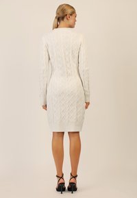 Weißes gestricktes Kleid mit einem strukturierten Rautenmuster, langen Ärmeln, geripptem Saum und figurbetonter Silhouette, kombiniert mit schwarzen Riemchen-High-Heels.