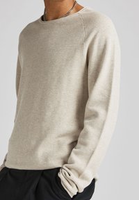 Pull à manches longues beige clair avec un motif tricoté texturé, col rond et poignets retroussés, porté avec un pantalon foncé.