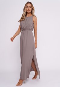 Langes, ärmelloses, taupefarbenes Kleid mit taillierter Taille und seitlichem Schlitz, aus glattem Stoff mit leichtem Glanz. Sichtbare, hautfarbene Sandalen.