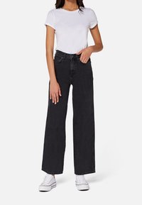 Žena oblečená v bílem krátkém tričku s krátkým rukávem, černých vysokých kalhotách s širokými nohavicemi a bílých platformových teniskách, stojí se jednou rukou v kapse.