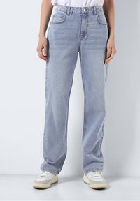 Person klädd i ljusblå raka jeans, vita sneakers och en vit skjorta, som står mot en enkel grå bakgrund.