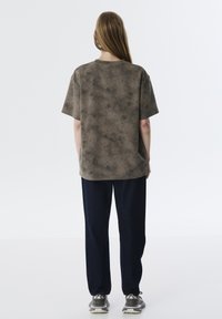 Korte mouwen, losvallend T-shirt in een gemêleerde bruine stof, gecombineerd met donkerblauwe broeken en grijze sneakers, bekeken vanaf de achterkant.