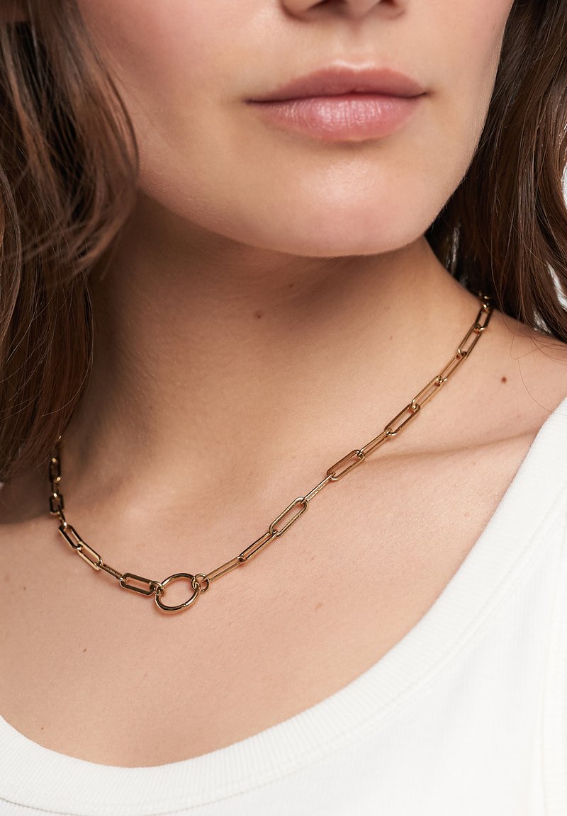 Goldfarbene Metallkettenglieder mit länglichen Büroklammer-ähnlichen Links und einem runden Verschluss, präsentiert auf einem hellen Oberteil.