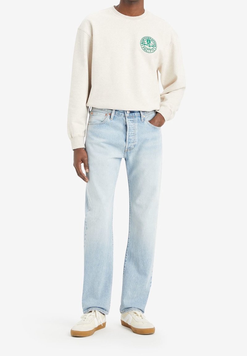 Sudadera de cuello redondo color beige claro con un logotipo gráfico verde en el pecho, combinada con jeans rectos de color azul claro y zapatillas blancas.