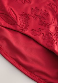 Tissu en satin rouge présentant des motifs ton sur ton de feuilles et de fleurs brodées, avec une texture lisse et des bords soigneusement cousus.