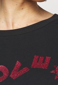 Černé bavlněné tričko s širokým výstřihem, které nese červený třpytivý text, čímž vytváří texturovaný kontrast s hladkým materiálem.