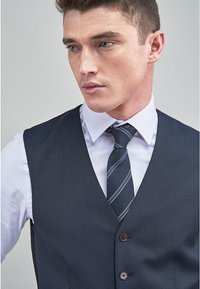 Gilet marine foncé à coupe ajustée, doté de trois boutons et d'une texture lisse, associé à une chemise habillée claire et une cravate rayée.