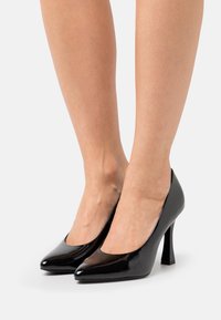 Zapatos de tacón alto de charol negro con punta afilada, textura suave y diseño de tacón cuadrado. Sin patrones o acentos visibles.