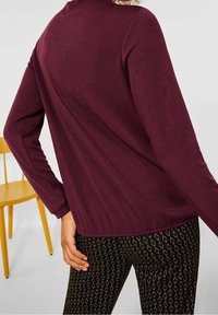 Burgunderfarbendes Langarmoberteil mit lockerer Passform, kombiniert mit gemusterten schwarzen Leggings mit geometrischem Design. Der Hintergrund zeigt einen gelben Stuhl.