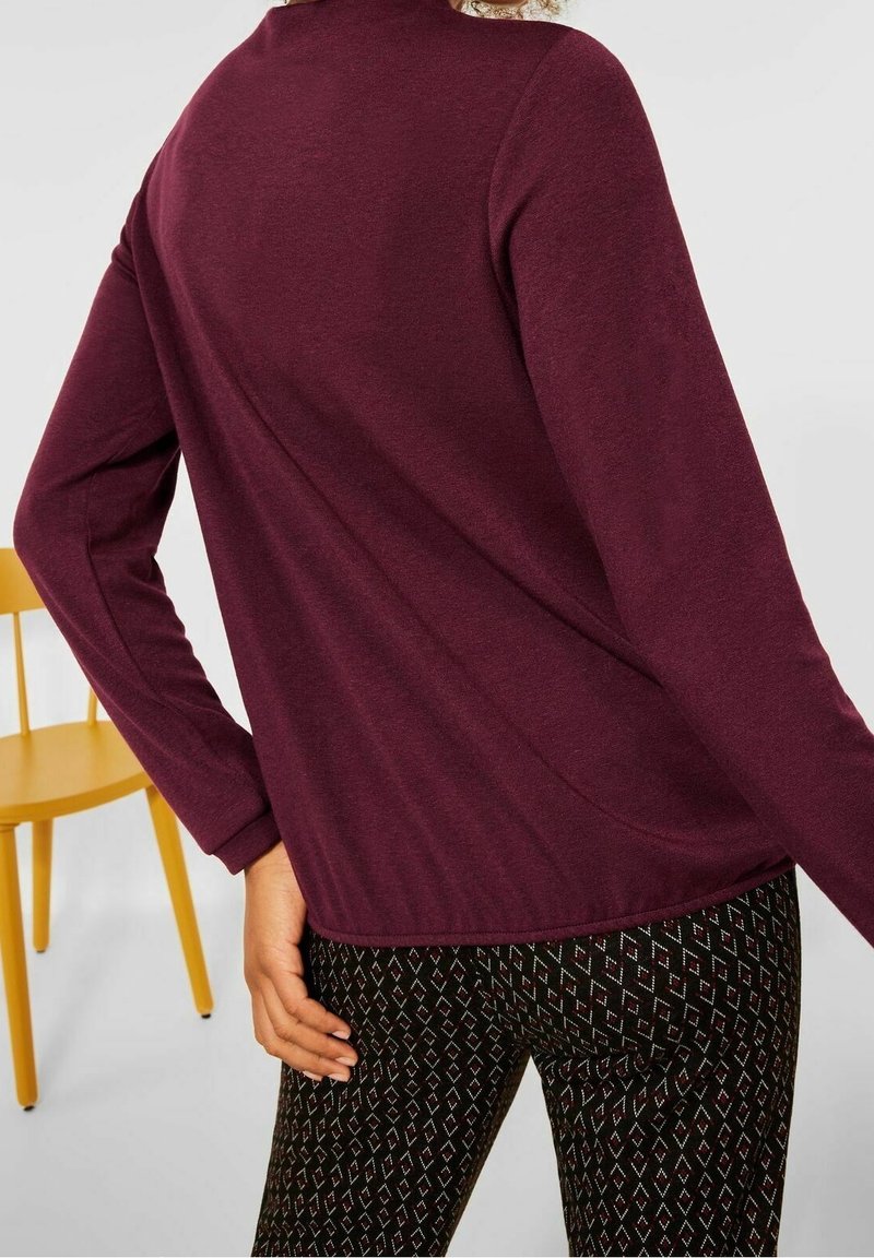 Burgunderfarbendes Langarmoberteil mit lockerer Passform, kombiniert mit gemusterten schwarzen Leggings mit geometrischem Design. Der Hintergrund zeigt einen gelben Stuhl.