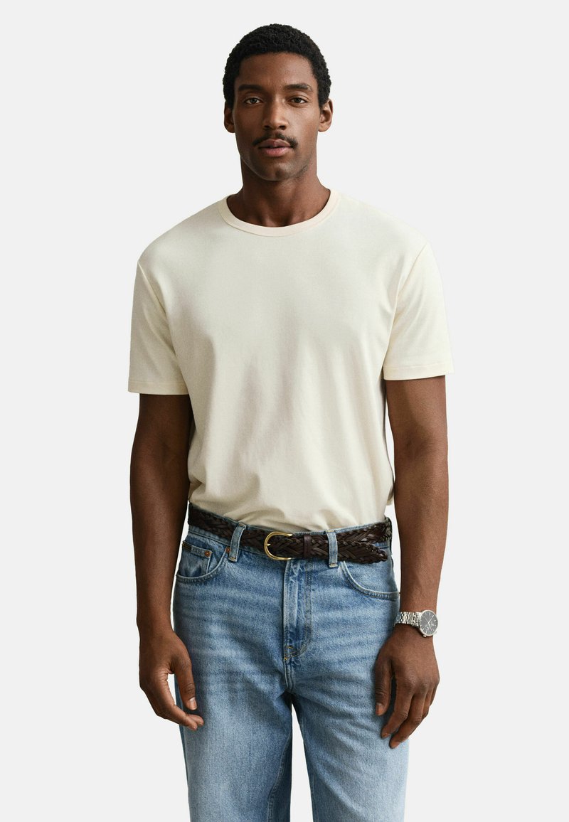 Homme portant un t-shirt uni blanc cassé, un jean bleu clair avec une ceinture tressée marron et une montre-bracelet argentée, debout devant un fond blanc.
