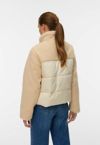 Sieviete ar brūnu zirgasti, tērpusies bēša teksturēta vilnas jaciņā ar pildītu aizmugures paneli un zilām džinsu biksēm, skatās prom uz balta fona.