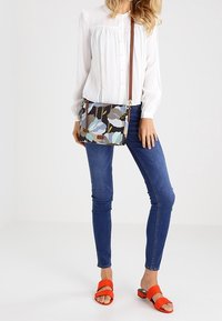 Kwiatowa torba na ramię z brązowo-niebieskim wzorem, prostokątnym kształtem, czarnym akcentem i regulowanym brązowym paskiem. Noszona z białą bluzką i dżinsami.