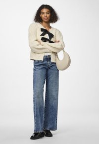 Κρεμ πλεκτό ζακέτα με μαύρες κορδέλες, σε συνδυασμό με ψηλόμεσες φαρδιές τζιν μπλε και μαύρες πατέντες loafers. Κρατάει μια μπεζ τσάντα.