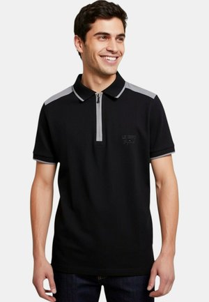 Jeune homme souriant, portant un polo noir avec une fermeture éclair grise et des accents aux épaules, debout devant un fond blanc uni.