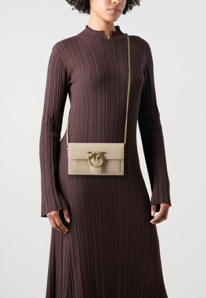 Borsa a tracolla beige con chiusura ad anello in tonalità oro e dettaglio a forma di uccello. Il materiale è pelle liscia. Il modello indossa un vestito a coste scuro.