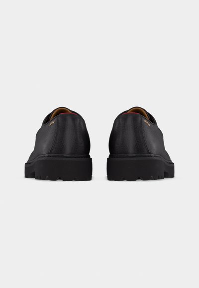 Paire de chaussures habillées en cuir noir avec semelles épaisses à crampons, coutures visibles et petit logo doré "HUGO" près du talon, vues de dos.