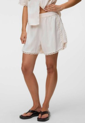 Kvinde iført hvide løstsiddende shorts med bølget blondekant og sorte platforme flip-flops, stående med den ene hånd på hoften og den anden afslappet.