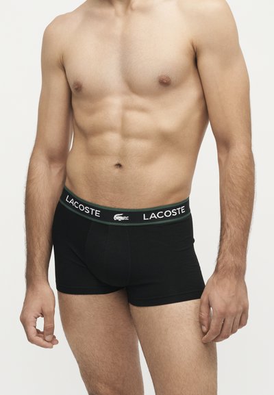 Homme torse nu portant un boxer noir Lacoste avec une ceinture logo, debout devant un fond clair uni.