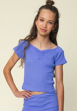 Tienermeisje met lang haar in een knot, gekleed in een blauw geribd kortärmelig topje en bijpassende rok, staand met een hand op de heup tegen een neutrale achtergrond.