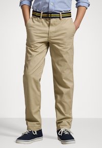 Garçon portant un chino beige avec une ceinture rayée noire et jaune, des baskets en toile navy et une chemise à rayures bleues à manches longues retroussées.
