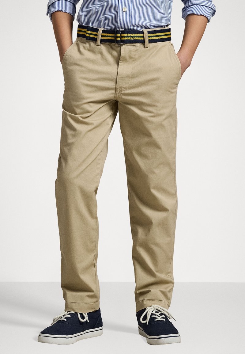 Garçon portant un chino beige avec une ceinture rayée noire et jaune, des baskets en toile navy et une chemise à rayures bleues à manches longues retroussées.