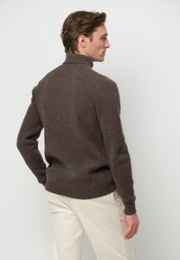 Brauner gerippter Strickpullover mit hohem Kragen, langen Ärmeln und taillierter Silhouette. Getragen über hellfarbigen Hosen.