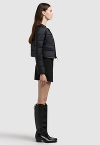 Kvinde i sort cropped dunjakke, sorte shorts og høje sorte læderstøvler stående i profil mod en ensfarvet baggrund.