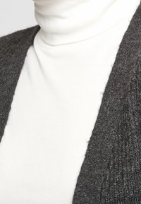 Grijze gebreide cardigan over een witte coltrui. De cardigan heeft een textuurpatroon en een open voorkant.