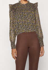 Blusa floreale con spalle a balza. Sfondo nero con fiori rosa e gialli. Tessuto trasparente, abbinata a pantaloni a coste marroni.