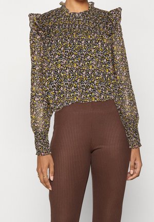 Blusa floreale con spalle a balza. Sfondo nero con fiori rosa e gialli. Tessuto trasparente, abbinata a pantaloni a coste marroni.