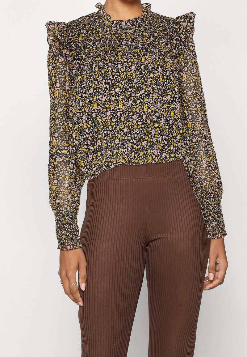 Blusa floreale con spalle a balza. Sfondo nero con fiori rosa e gialli. Tessuto trasparente, abbinata a pantaloni a coste marroni.