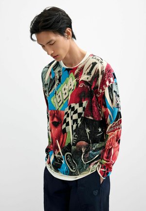 Giovane con capelli scuri guarda verso il basso, indossa una camicia colorata a maniche lunghe con stampe grafiche che includono "1984" e bandiere a scacchi.