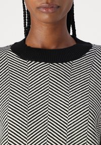 Suéter listrado em preto e branco com um colarinho liso preto e uma textura padrão com design de espinha de peixe. Vista em close.