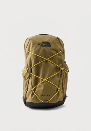 Mochila North Face Jester en verde oliva con base negra, cordones elásticos amarillos en el frente, bolsillos laterales de malla y cierre superior con cremallera.