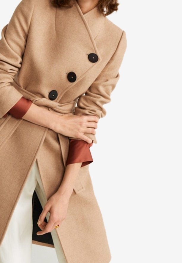 Femme portant un manteau beige croisé avec des boutons noirs, des poignets de chemise couleur rouille et un pantalon blanc, les bras croisés devant elle.