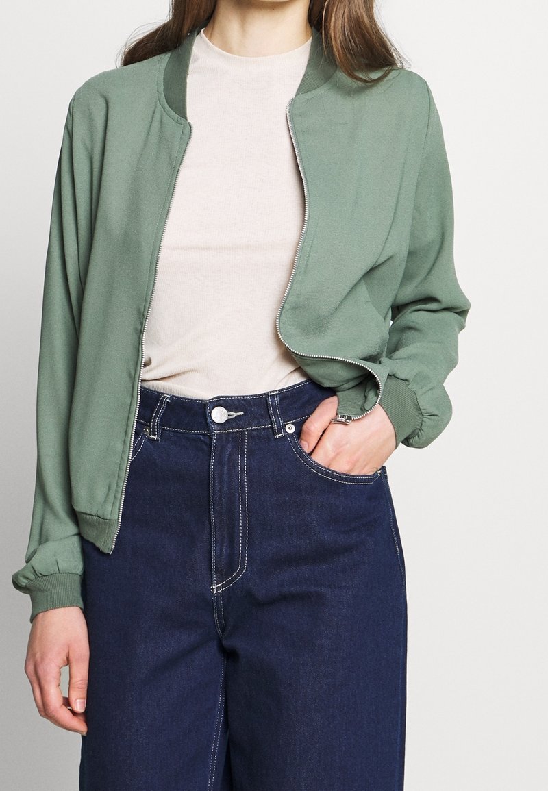 Zelená bomber bunda se zipem, žebrovanými manžetami, lehkým materiálem, nošená přes světle šedý top, spárovaná s tmavě modrými džíny.