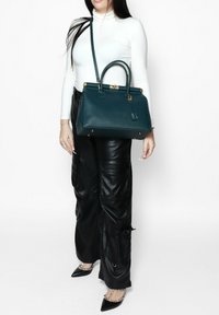 Teal leren handtas met gestructureerde vorm, gouden hardware, bovenhandvatten en een afneembare schouderriem. Draagt door een persoon in een aansluitend wit topje en zwarte broek.