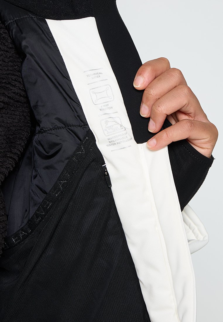 Giacca nera in tessuto esterno con fodera interna bianca, dotata di una tasca con zipper e etichette di istruzioni per la cura su un lato. Texture morbida visibile.