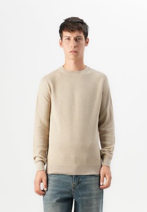 Joven de pie que lleva un suéter de punto texturizado beige y jeans azules frente a un fondo blanco liso.