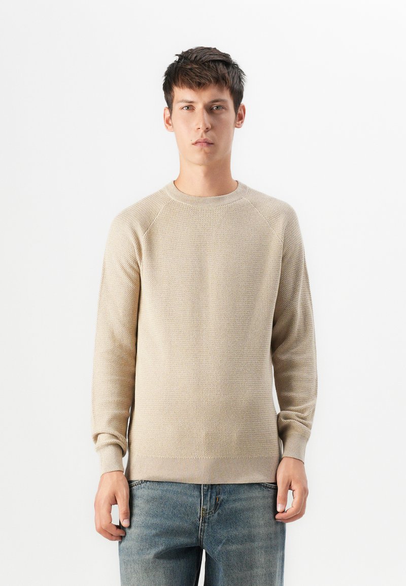 Jeune homme debout portant un pull à mailles texturées beige et un jean bleu devant un fond blanc uni.