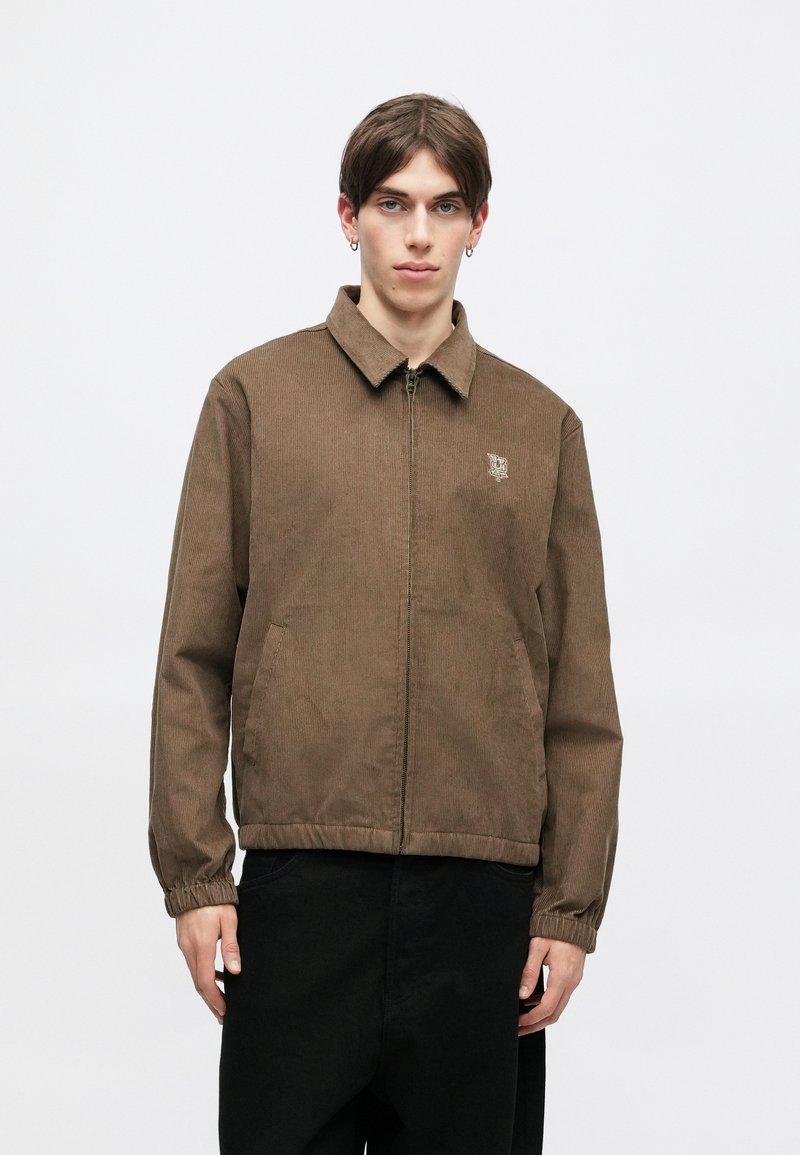 Brun kordfløyelsjakke med krage, sidelommer og elastiske ermene. Gylden logo detalj på brystet. Brukt over svarte bukser.