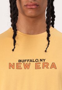 Žuta pamučna majica s kratkim rukavima koja sadrži tekst "BUFFALO.NY" i "NEW ERA" u kontrastnim crnim i narančastim bojama. Okrugli izrez.