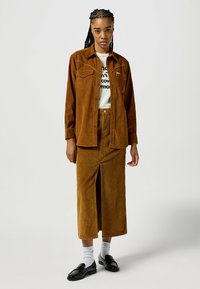 Chemise en velours côtelé marron avec des boutons-pression, associée à une jupe longue assortie avec une fente à l'avant. Des mocassins noirs et des chaussettes blanches complètent la tenue.